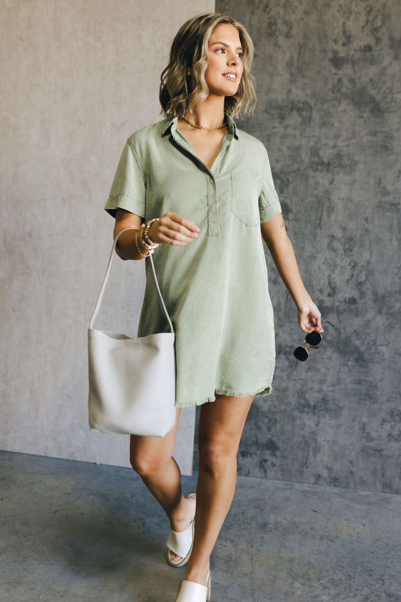 Full body view of model wearing the Aubrie Washed Olive Short Sleeve Mini Dress which features washed olive tencel fabric, one front chest pocket, a hidden quarter button-up, a v neckline with a collar, a frayed mini-length hem, and cuffed short sleeves.