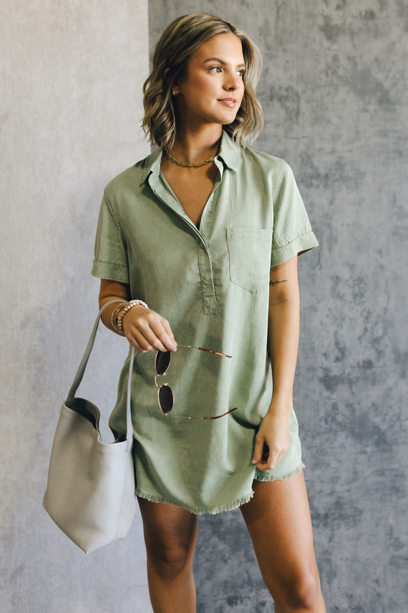 Front view of model wearing the Aubrie Washed Olive Short Sleeve Mini Dress which features washed olive tencel fabric, one front chest pocket, a hidden quarter button-up, a v neckline with a collar, a frayed mini-length hem, and cuffed short sleeves.