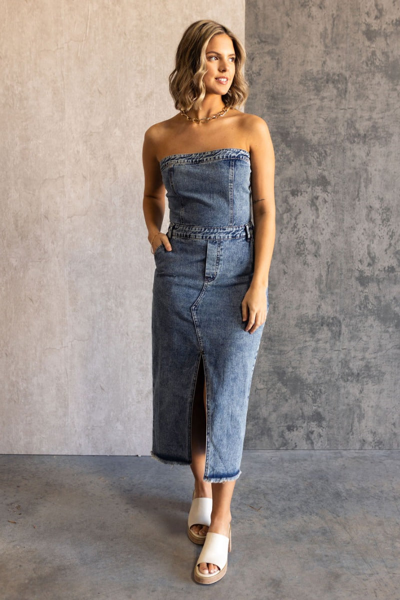 Full body view of model wearing the Carrie Washed Denim Slit Strapless Midi Dress which features medium denim wash fabric, brown stitch details, front slit detail, midi length, two front slit pockets, two back pockets, belt loops, frayed hem, straight acr