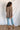 Full body back view of model wearing the Easy Breezy Top, that has a mocha colored knit material, an open knit design, a V neckline, a long cuffed sleeve, side slits, and a high-low design.
