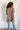 Back view of model wearing the Easy Breezy Top, that has a mocha colored knit material, an open knit design, a V neckline, a long cuffed sleeve, side slits, and a high-low design.