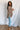 Full body side view of model wearing the Easy Breezy Top, that has a mocha colored knit material, an open knit design, a V neckline, a long cuffed sleeve, side slits, and a high-low design.