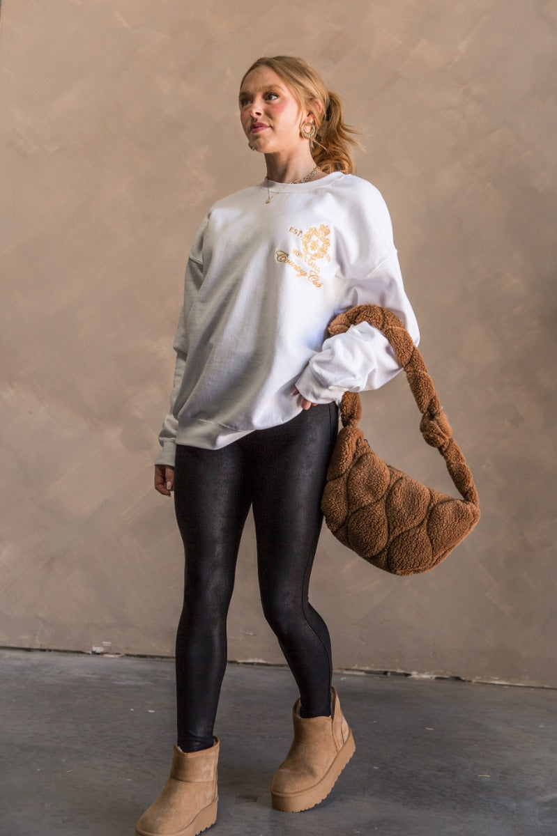 Full body view of model wearing the Monte Carlo White Long Sleeve Sweatshirt which features white knit fabric, thick hem, round neckline, golden stitched design says "EST. 1974 MONTE CARLO Country CLub" with gold crown and long sleeves with cuffs.