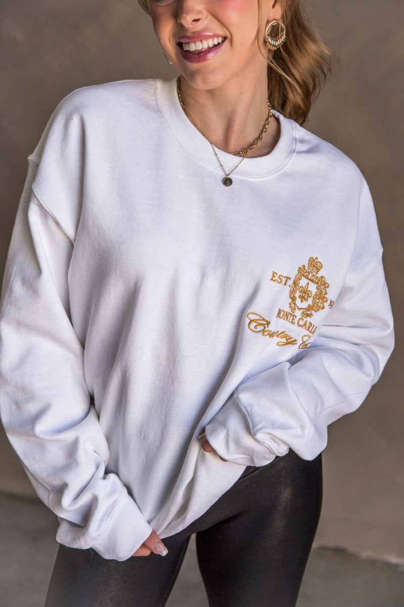 Front view of model wearing the Monte Carlo White Long Sleeve Sweatshirt which features white knit fabric, thick hem, round neckline, golden stitched design says "EST. 1974 MONTE CARLO Country CLub" with gold crown and long sleeves with cuffs.