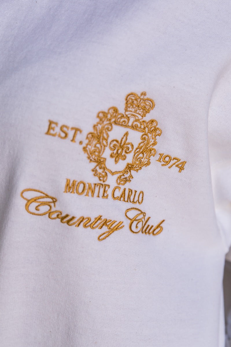 Clos eup view of model wearing the Monte Carlo White Long Sleeve Sweatshirt which features white knit fabric, thick hem, round neckline, golden stitched design says "EST. 1974 MONTE CARLO Country CLub" with gold crown and long sleeves with cuffs.