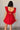 Back view of model wearing the Aurora Red Floral Ruffle Mini Dress which features red fabric with a brown 3d floral pattern, a mini-length baby doll style, a v neckline, elastic straps with ruffles, and an open back with a bow detail.