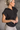 Side view of model wearing the Presley Black Sleeveless Hoodie Sweatshirt which features black knit fabric, monochrome quarter zip up, high neckline, hoodie attached with drawstring details, one front pocket and sleeveless.