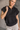 Front view of model wearing the Presley Black Sleeveless Hoodie Sweatshirt which features black knit fabric, monochrome quarter zip up, high neckline, hoodie attached with drawstring details, one front pocket and sleeveless.
