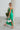 Side view of the Call You Mine Midi Dress In Green that features a green colored silk material, a cowl neckline, a sleeveless design with thin adjustable straps, a side slit, and a midi length.