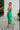 Side view of the Call You Mine Midi Dress In Green that features a green colored silk material, a cowl neckline, a sleeveless design with thin adjustable straps, a side slit, and a midi length.