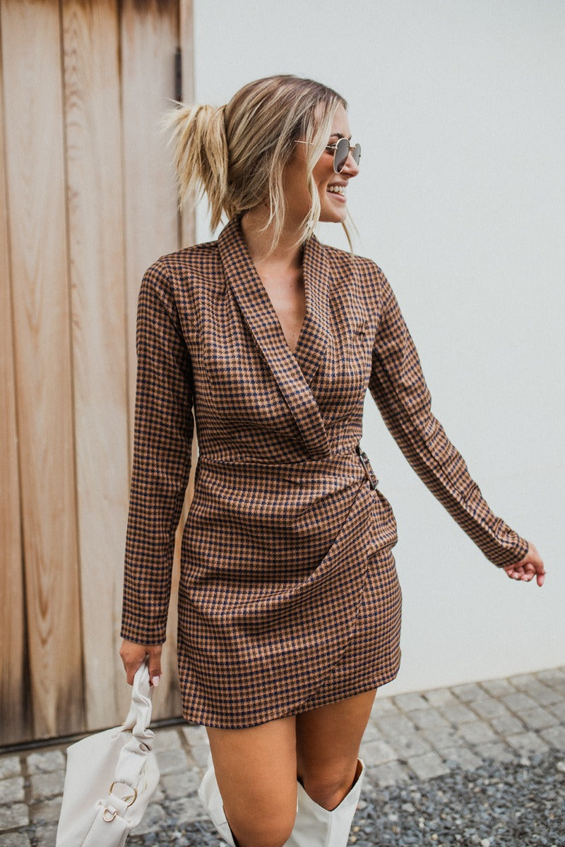 Frontal view of the Next Century Dress that features a brown tweed material, a navy gingham print, a lapel surplice neckline, a long sleeve, a wrap front with a tortoiseshell buckle, and a mini length.