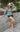 Full body front view of model wearing the Forever Young Top in Sage, which features a sage green colored silky material, a round neckline with a ruffle detail, a notched neckline with adjustable ties, long sleeves, and a flowy fit