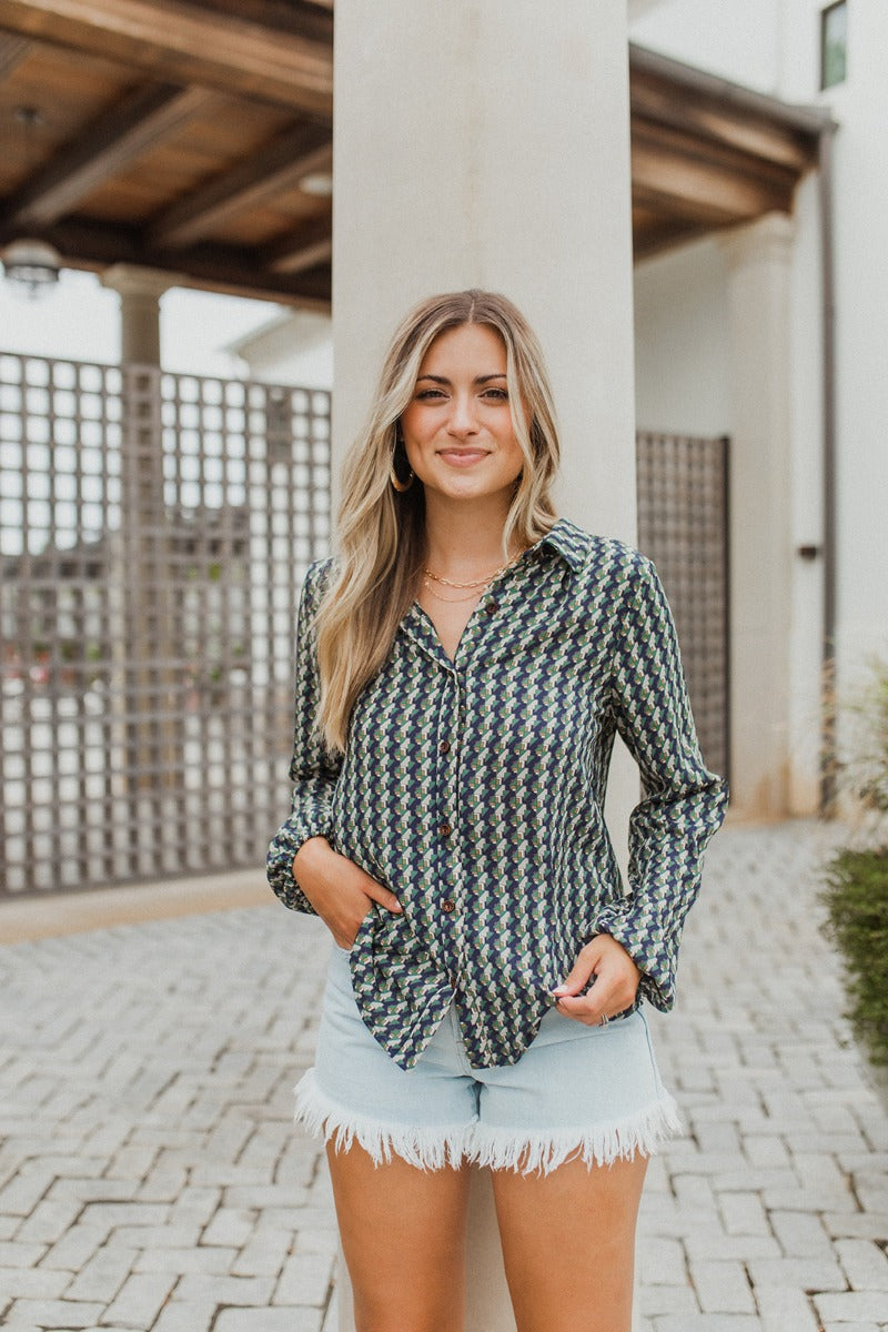 Frontal view of the Around The Block Top that features a navy chiffon material, a taupe and green geometric print, a collar neckline, a button-up front, and a long sleeve.
