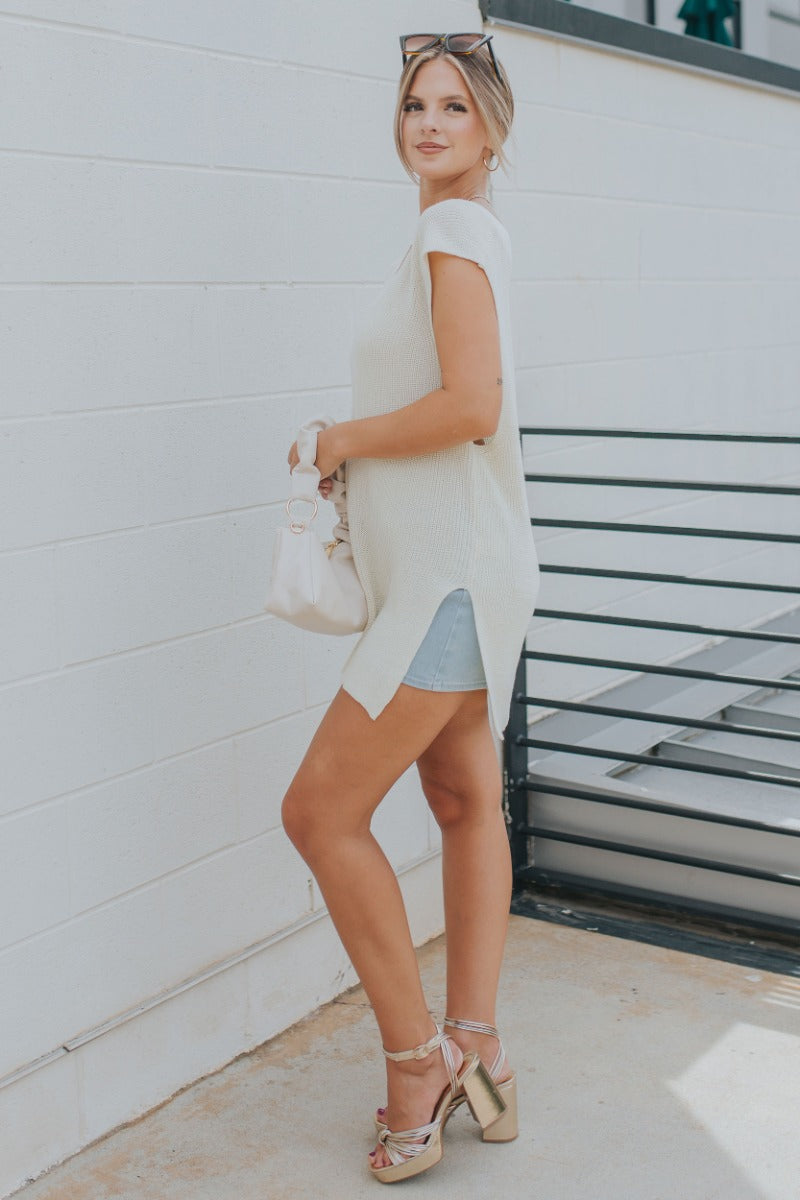 Full body side view of model wearing the It's Show Time Vest in Cream which features cream cable knit fabric, slits on each side, v-neckline and sleeveless.