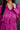 Close-up front view of model wearing the Dancing Queen Dress, that has a fuchsia textured material, a satin collar neck, a button-up front, long cuffed sleeves, an adjustable tie belt, and lettuce edge bottom hem.