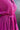 Close-up front/side view of model wearing the Dancing Queen Dress, that has a fuchsia textured material, a satin collar neck, a button-up front, long cuffed sleeves, an adjustable tie belt, and lettuce edge bottom hem.