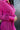 Close-up front/side view of model wearing the Dancing Queen Dress, that has a fuchsia textured material, a satin collar neck, a button-up front, long cuffed sleeves, an adjustable tie belt, and lettuce edge bottom hem.