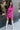 Full body front view of model wearing the Dancing Queen Dress, that has a fuchsia textured material, a satin collar neck, a button-up front, long cuffed sleeves, an adjustable tie belt, and lettuce edge bottom hem.
