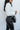 Close-up side view of 5'8 model wearing the Next Level Straight Leg Pants features a black faux-leather material, a high-rise fit, a clasp and zipper front closure, belt loops, two front pockets, a middle seam down the legs, and a straight leg fit