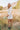 Full body side view of model wearing the Daydreamer Faux Leather Dress, that has an ivory PU material, a surplice neckline, shoulder pads, long sleeves, a draped front, a back zipper closure, and a mini length