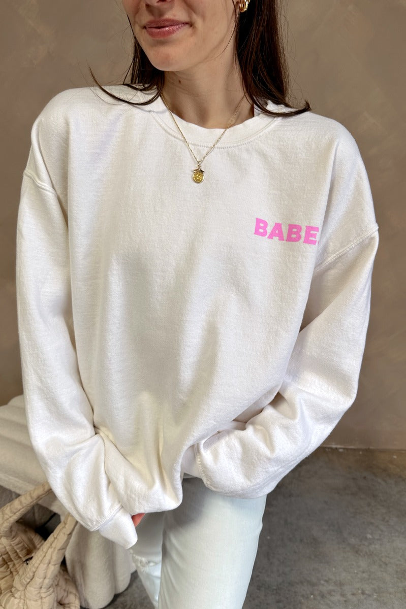 Front view of model wearing the Babe Pink & Cream Long Sleeve Sweatshirt which features cream knit fabric, thick hem, round neckline and long sleeves with cuffs. Graphic says "BABE" in pink with a mini heart design and "BABE" in pink letterman on the back