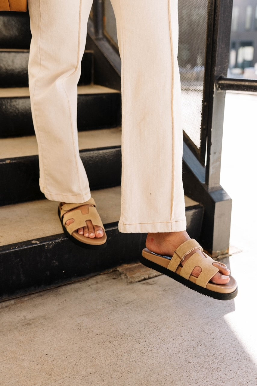 Front view of model wearing the Mayven Sandal in Taupe Suede which features taupe suede fabric, adjustable hook and velcro closure strap, "h" shaped strap, round toe, slide one style and black sole.