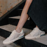 Front right view of female model wearing the Zina Crystal Sneaker in Ivory Suede which features ivory suede upper fabric, crystal stone design, white lace up closure, silver and neutral tones on sole and round toe.