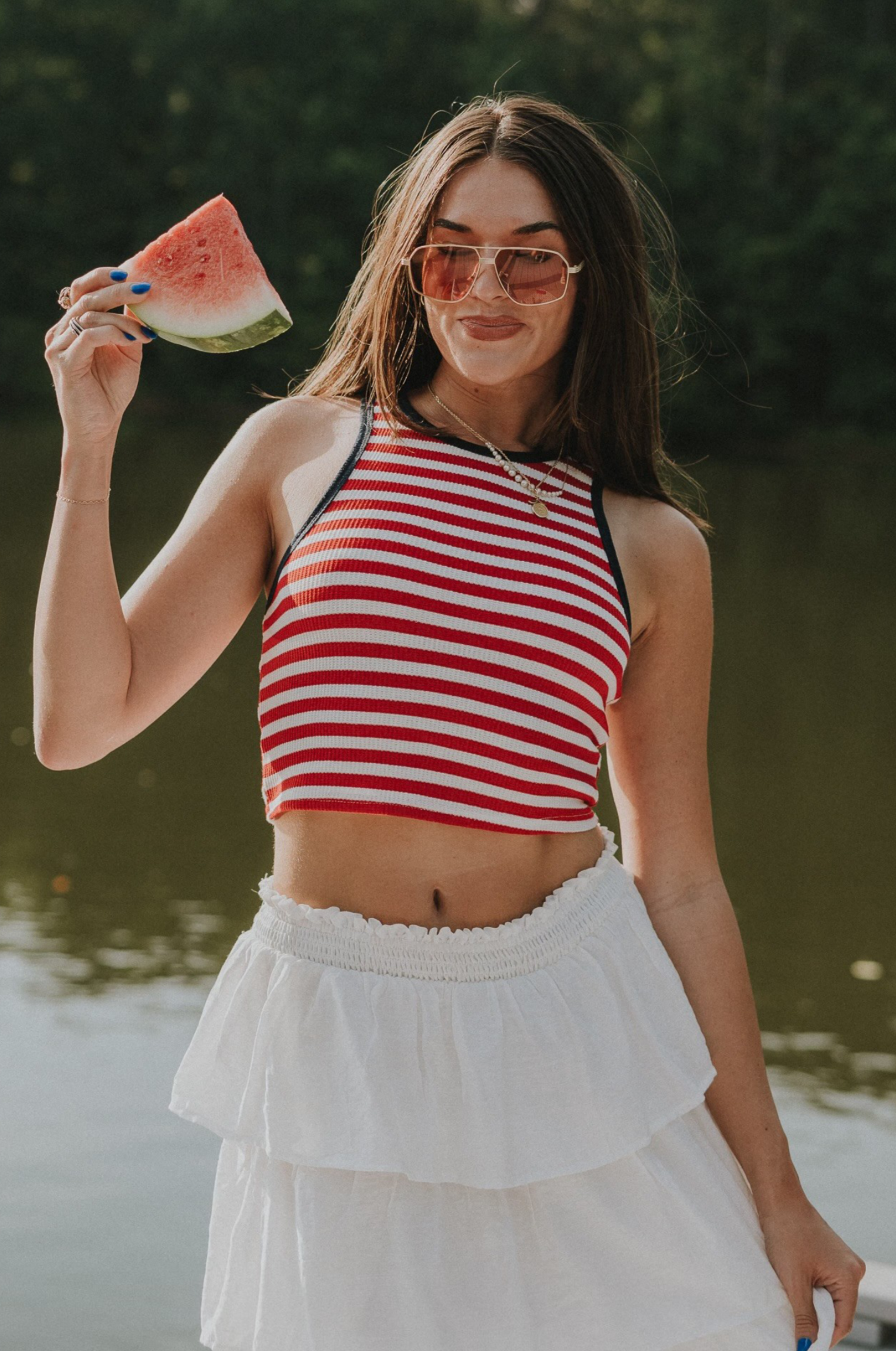 front view of female model wearing the Felicity Red, White, & Blue Striped Tank Top that has red and white horizontal stripes, navy trim, a cropped waist, and thick straps. Worn with white skirt.