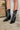Close up frontal view of a model wearing the Emersyn Western Boots, that feature black faux-leather with white western stitching, slip on entry, and 3.5" heels