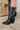 Close up left side view of a model wearing the Emersyn Western Boots, that feature black faux-leather with white western stitching, slip on entry, and 3.5" heels