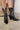 Close up front/side view of a model wearing the Emersyn Western Boots, that feature black faux-leather with white western stitching, slip on entry, and 3.5" heels