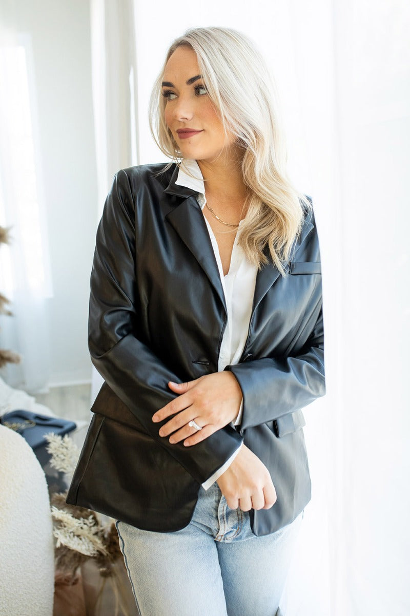 Front view of model wearing the Back To Business Blazer in Black that has  black faux-leather, a collar and lapel, and long sleeves. Worn over white blouse.