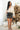 Full body front view of model wearing the Most Wanted Skirt in Black that has a black faux-leather material, a front overlay with ruching on the side, and a back zipper closure