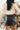 Close back view of model wearing the Most Wanted Skirt in Black that has a black faux-leather material, a front overlay with ruching on the side, and a back zipper closure
