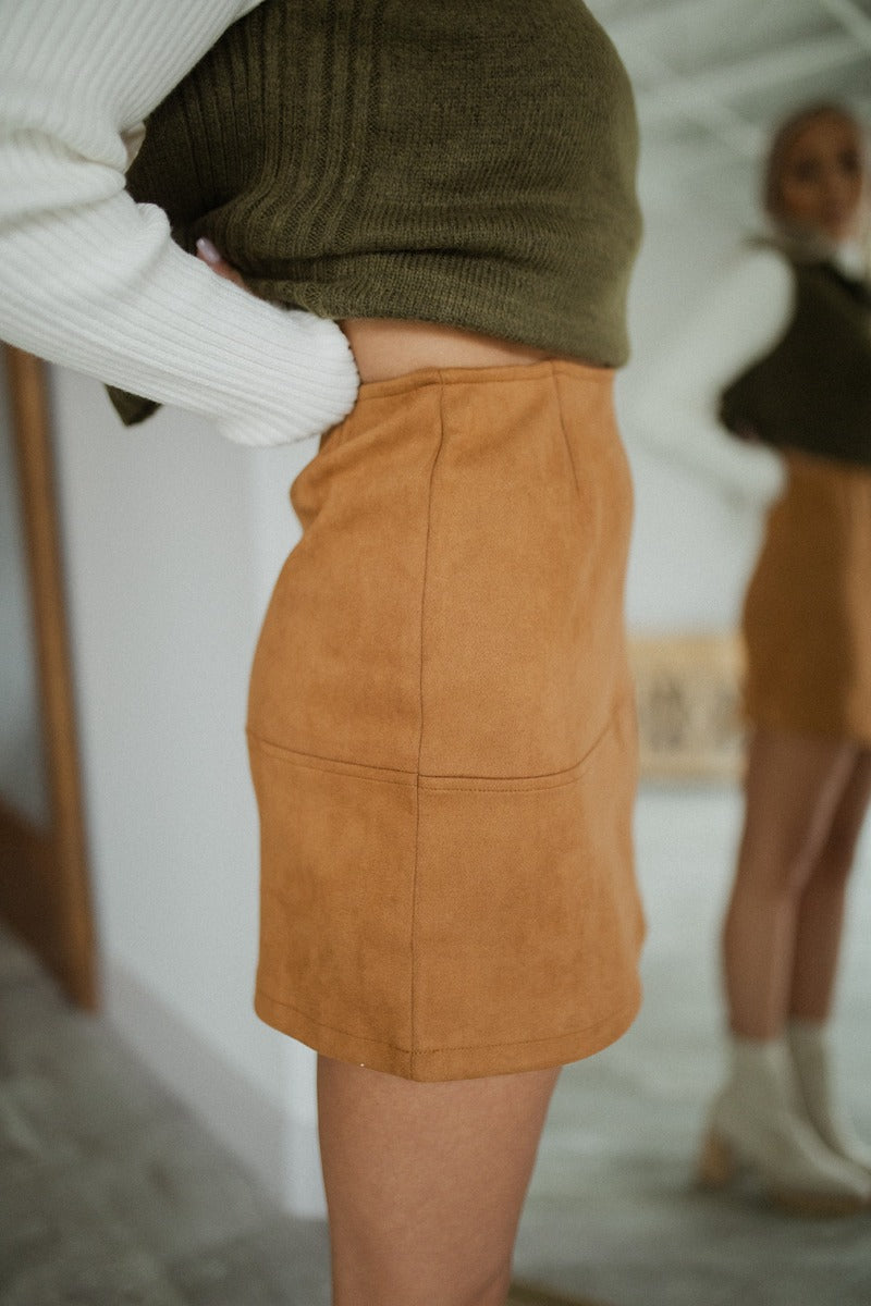 Side view of model wearing the Static Radio Skirt in Cognac that has cognac suede fabric, stitched details, a mini length hem, and an elastic waistband.