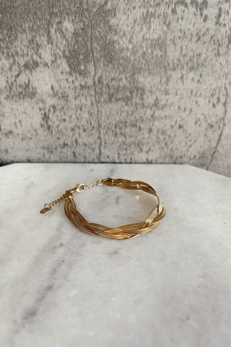 Top view of the Maeve Gold Braided Bracelet that has three gold flat bands braided together with an adjustable clasp closure shown against a marbled background.