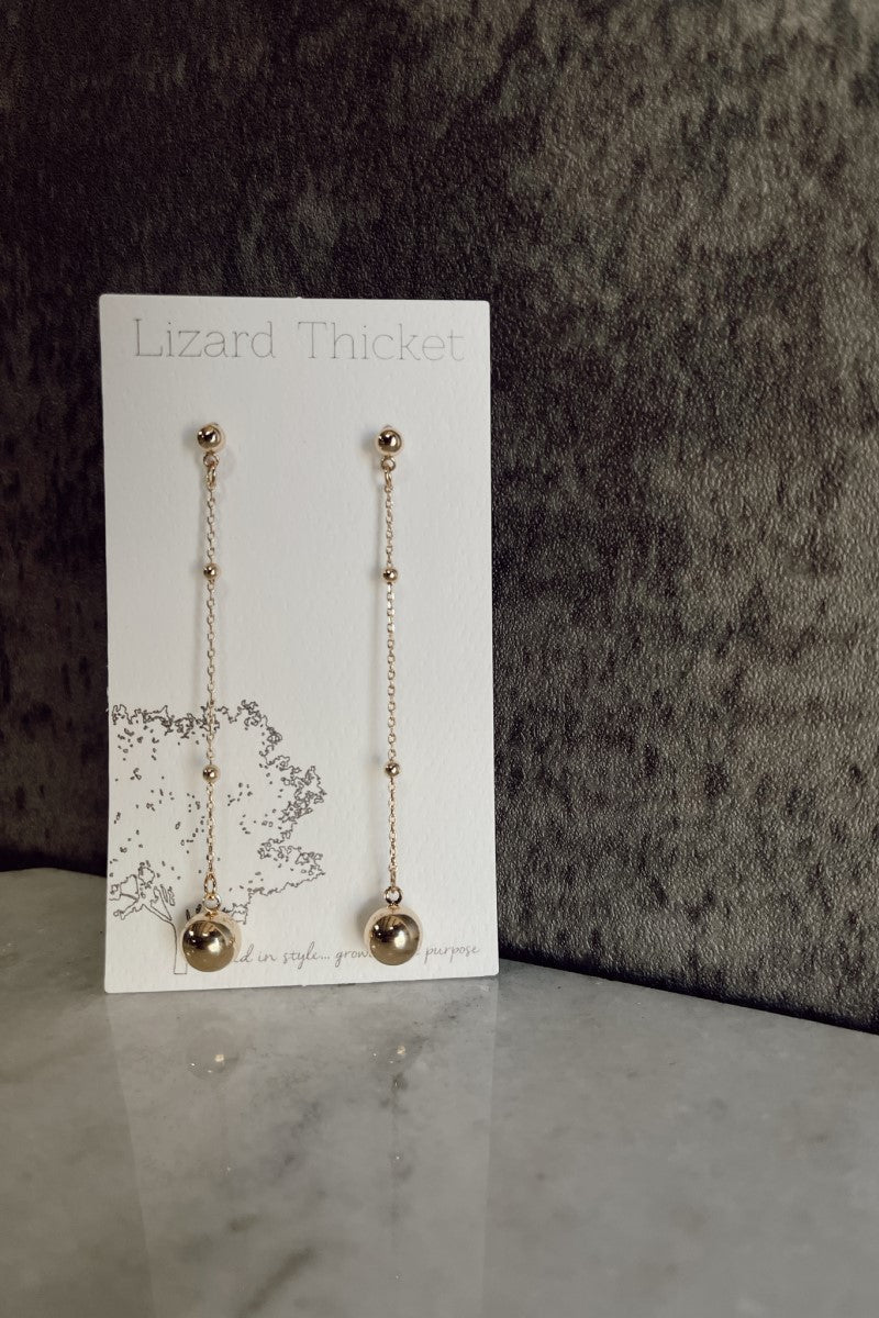 Image of the Isabel Gold Bead Drop Earrings that have gold dangle chains with gold ball beads and post backs, against a white earring card.