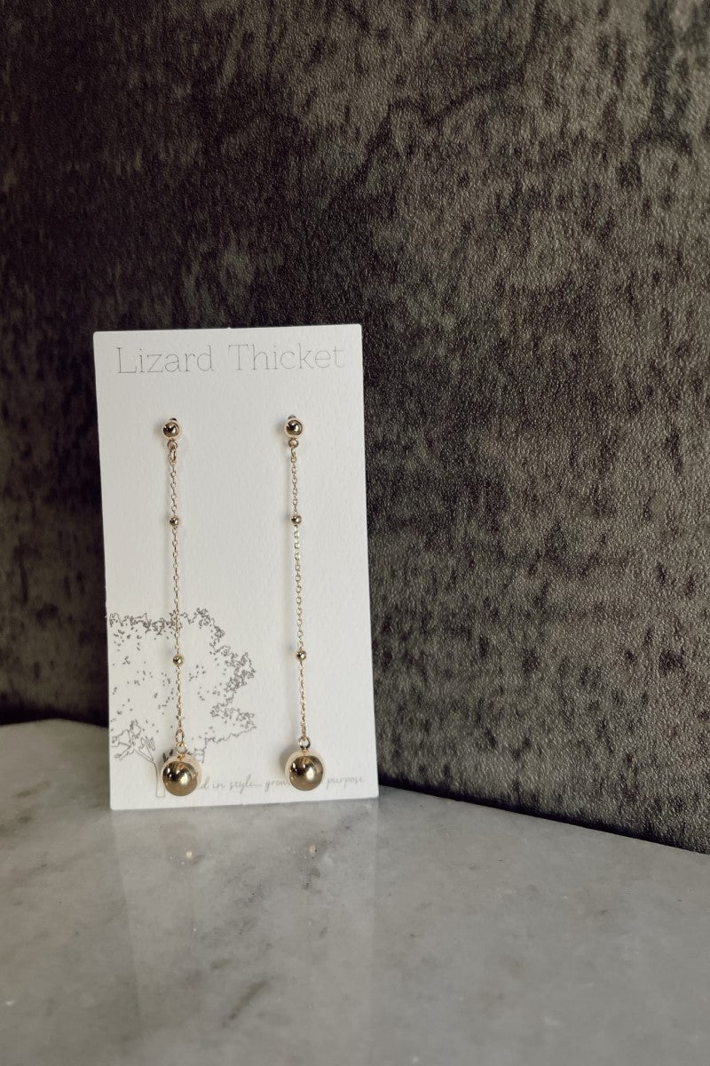 Image of the Isabel Gold Bead Drop Earrings that have gold dangle chains with gold ball beads and post backs, against a white earring card.