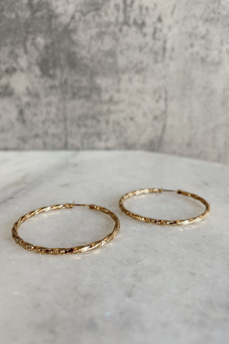Top view of the Ayla Gold Rope Hoop Earrings that have medium gold roped hoops with latch closures against a marbled background.