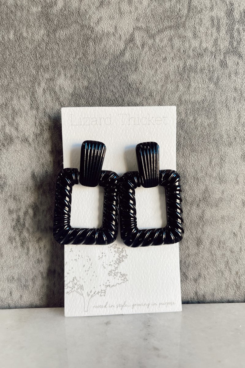 Front view of the Alina Black Earrings that have shiny black open rectangles with rope detailing and studs attached, pictured on white earring card.
