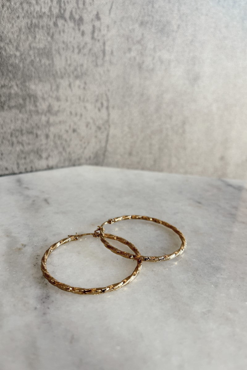 Top view of the Ayla Gold Rope Hoop Earrings that have medium gold roped hoops with latch closures against a marbled background.