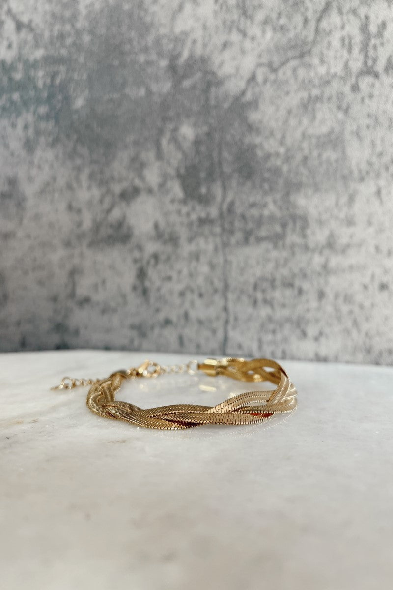 Front view of the Maeve Gold Braided Bracelet that has three gold flat bands braided together with an adjustable clasp closure shown against a marbled background.