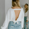 Back view of model wearing the Say No More Sweater, that features off white knit fabric with a stitched stripe pattern, a v neckline, an open back with lower back tie, and long balloon sleeves.