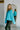 Side view of model wearing the Cool Air Sweater, that features turquoise knit fabric, a high neckline, and long balloon sleeves with cuffed wrists.