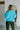 Back view of model wearing the Cool Air Sweater, that features turquoise knit fabric, a high neckline, and long balloon sleeves with cuffed wrists.