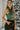 Side view of model wearing the Blaire Faux-Leather Bodysuit in Green that has green faux leather, a round neckline, a back button closure, a sleeveless body, and a knit thong bottom with snap closures. Worn with black pants.