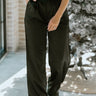 Front view of model wearing the Real Deal Satin Pants in Black that have black satin fabric, two front pockets, two back pockets, a front zipper and black button closure, belt loops, pleated detailing, and wide legs.