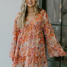 Front view of model wearing the Retro Revival Dress that has rust, teal and orange fabric with a paisley pattern, a ruffled hem, a v neckline, a lined body, a back button closure, and flare long sleeves