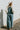 Back view of model wearing the One Way Ticket Overalls that feature dusty blue corduroy fabric, wide legs, one front pocket, two back pockets and button strap closures.