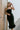 Front view of model wearing the The Big Dreams Midi Dress that has midi length black fabric, corset and ruched detailing, a feathered hem, s small slit in the back, a sleeveless body, and a lining.
